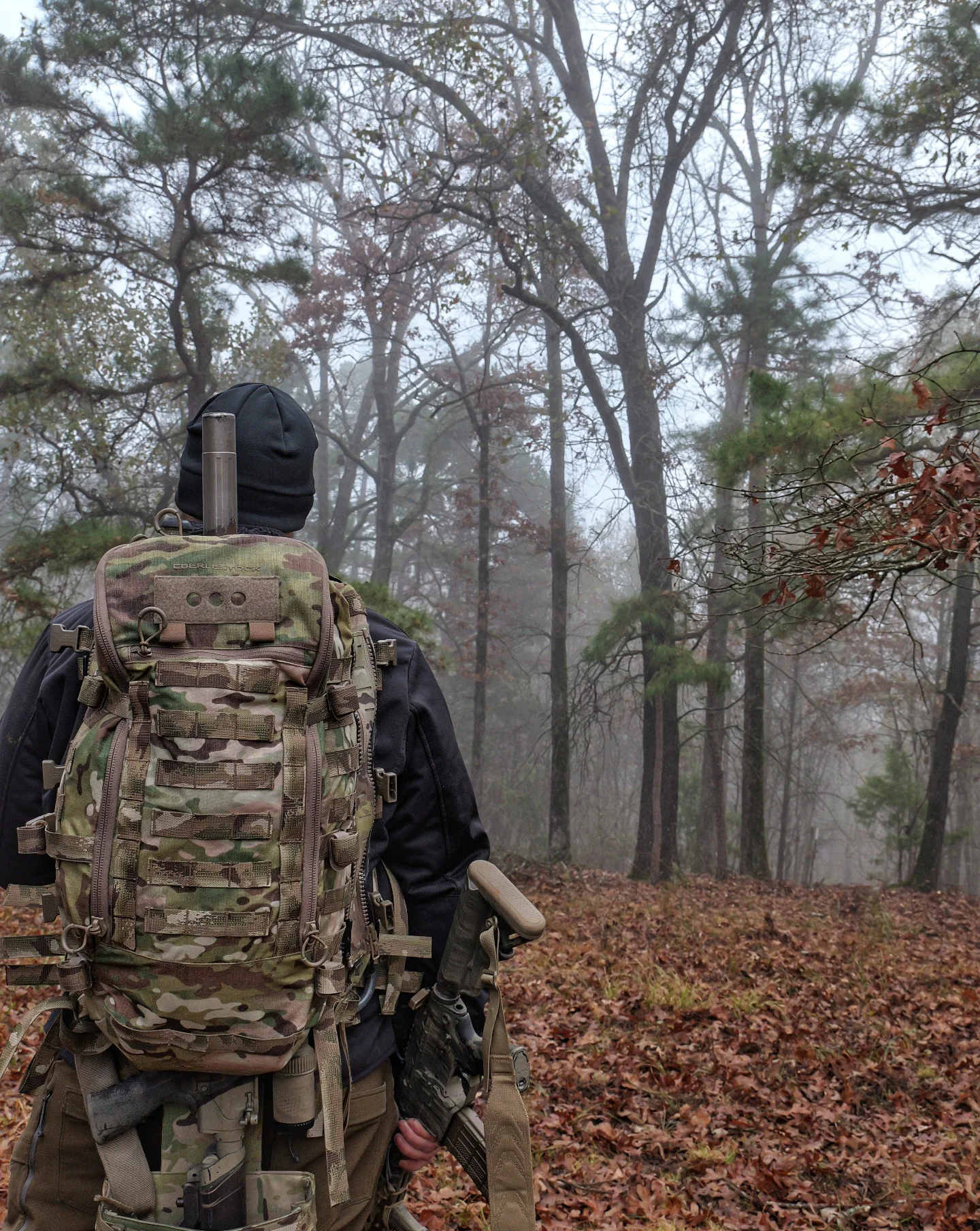 Rear view of hunter with Eberlestock pack and suppressed rifle facing misted timber